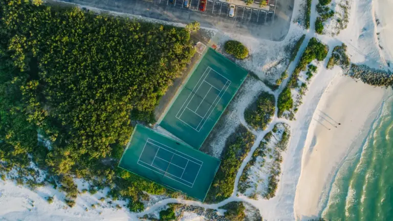 Vogelperspektive auf zwei Tennisplätze direkt am sandigen Strand neben einem Parkplatz.