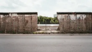Ein grauer Metallzaun vor den verbliebenen Überresten der Berliner Mauer bei Tageslicht.