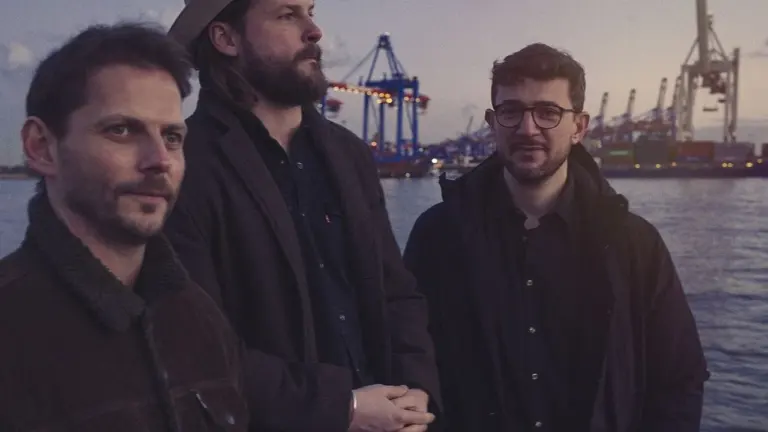 Drei Männer stehen vor einer Hamburger Hafen-Kulisse mit Kränen bei einsetzender Dämmerung im Freien.
