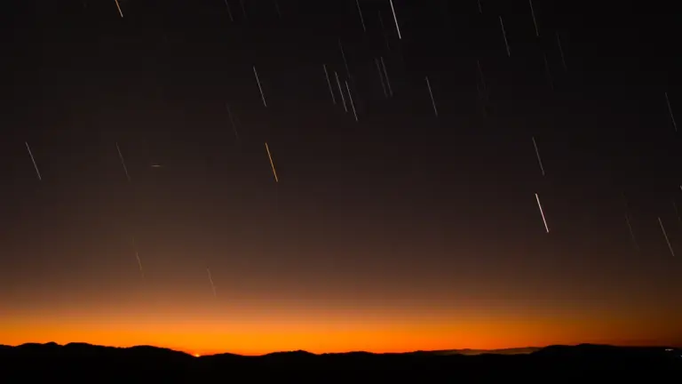 Lange Belichtungsaufnahme eines nächtlichen Sternenhimmels mit sichtbaren Lichtspuren über einer dunklen Hügellandschaft.
