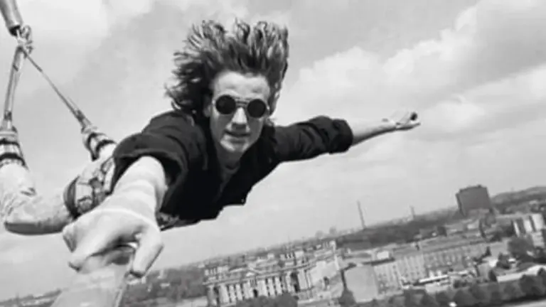 Ein junger Mann mit Sonnenbrille springt kopfüber beim Bungee-Jumping vor einer städtischen Skyline in die Tiefe.