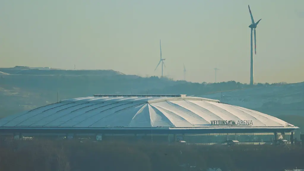 Die Veltins-Arena in Gelsenkirchen mit Windrädern im Hintergrund bei leichtem Tageslicht.