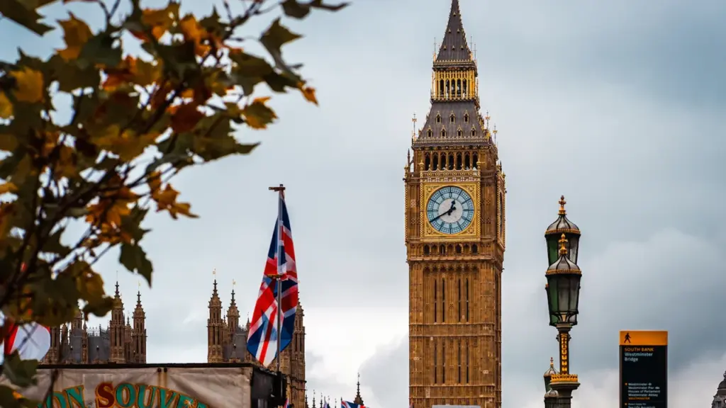 Big Ben Uhrturm in London unter einem bewölkten Himmel mit herbstlichen Blättern im Vordergrund.