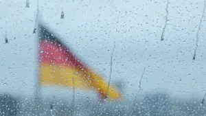 Die deutsche Flagge hinter einer verregneten Fensterscheibe mit vielen Wassertropfen bei grauem Wetter.