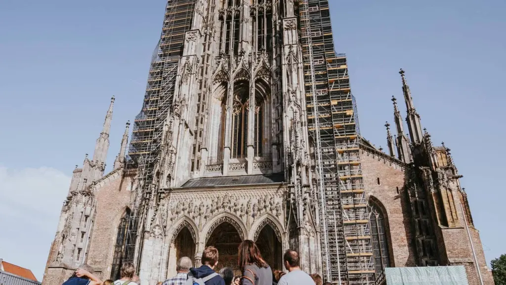 Eine Touristengruppe steht vor dem hohen Ulmer Münster mit Baugerüsten an der Fassade.