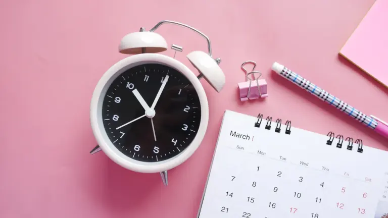 Weißer Wecker und Kalender auf rosa Hintergrund symbolisieren Zeitdruck und Termine.